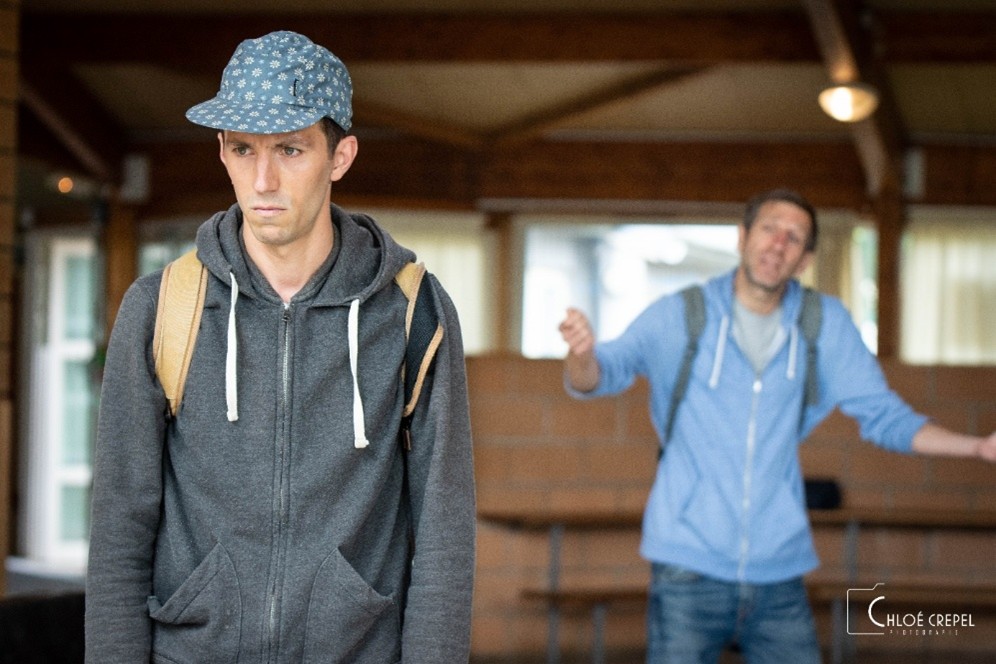 Un comédien fronce les sourcils aux premier plan, un comédien s'agite à l'arrière plan